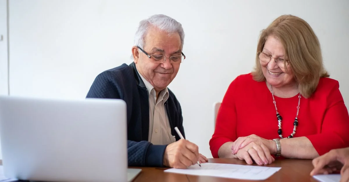 Couple âgé signant des documents à un bureau avec un ordinateur portable. Sourire et travail.