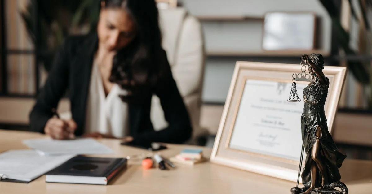 Femme concentrée écrivant à un bureau avec une figurine de la Justice et un certificat dans un bureau.