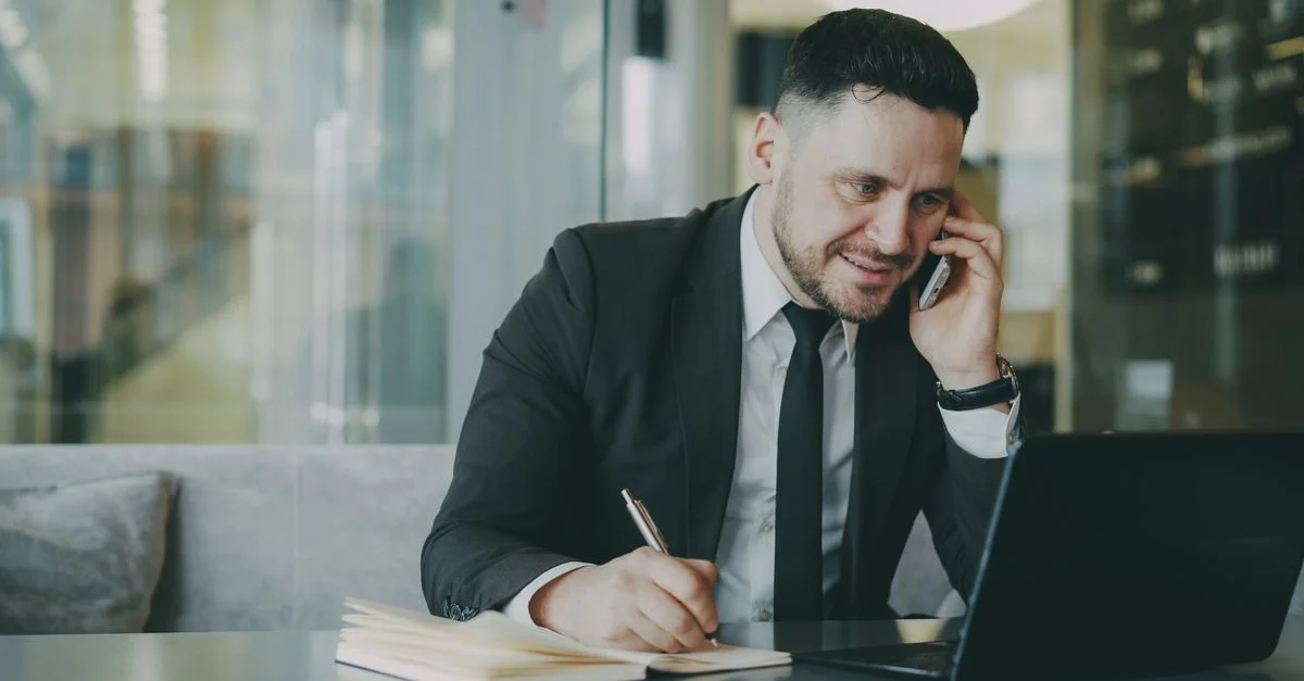 Un homme professionnel en costume jongle entre son téléphone et son ordinateur portable dans un bureau moderne.