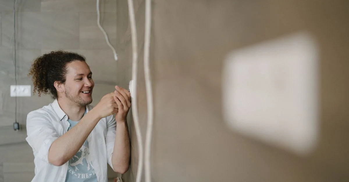 Un électricien avec des cheveux bouclés ajuste une prise à l'intérieur, souriant pendant qu'il travaille.