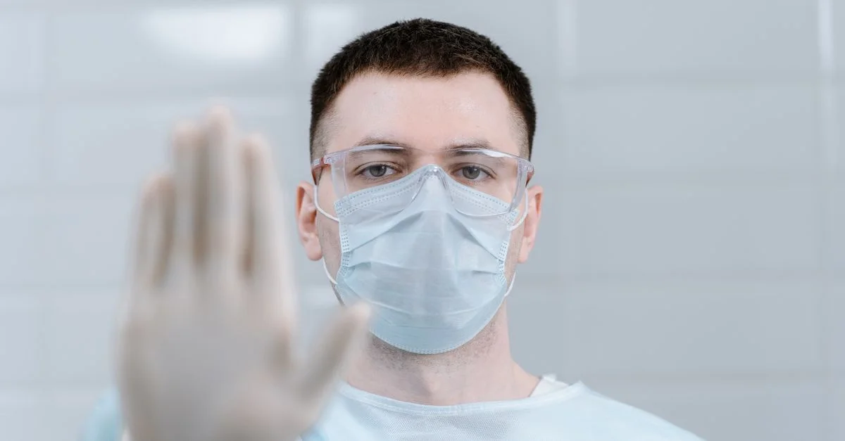 Travailleur de la santé portant un équipement de protection et un masque faisant un geste d'arrêt dans un cadre hospitalier.