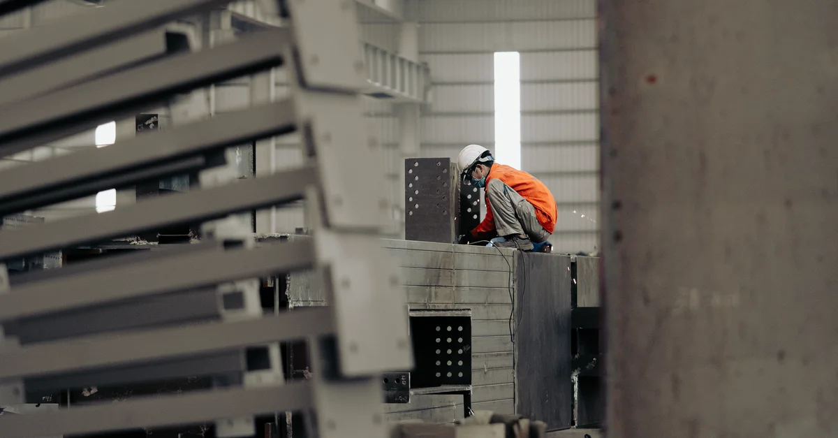 Ouvrier en uniforme de sécurité orange et casque blanc inspectant des structures métalliques dans une usine.