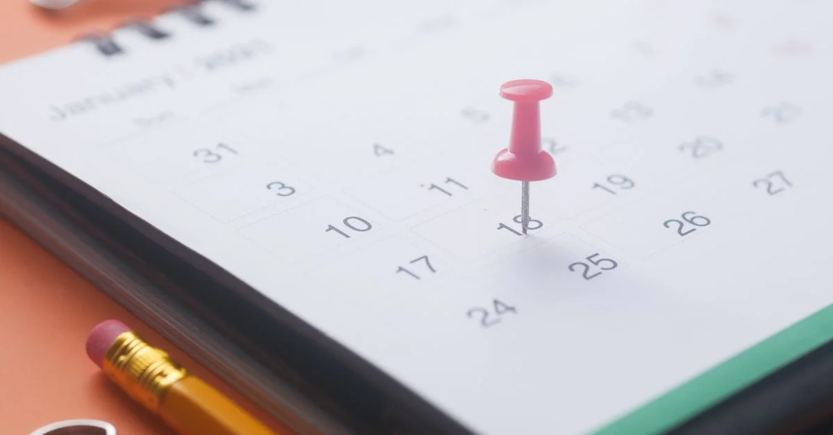 Gros plan d'une page de calendrier avec une épingle rose, un crayon et un clip sur un fond orange.
