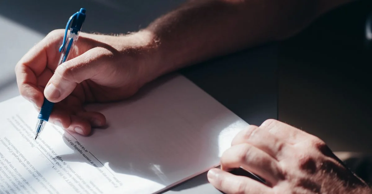 Une image en gros plan de la main d'une personne signant un document avec un stylo bleu dans un éclairage naturel.