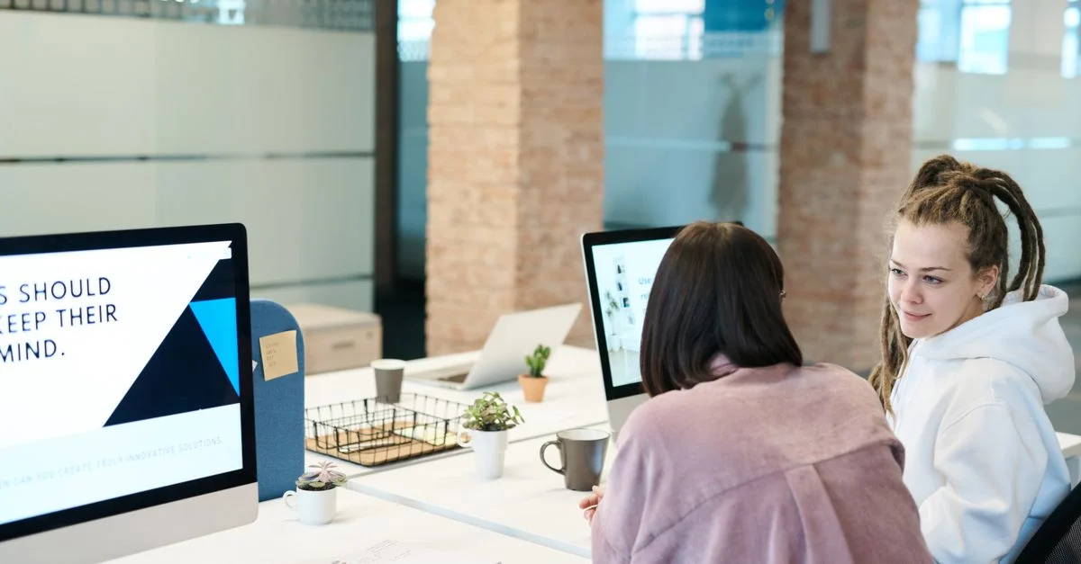 Deux femmes aux coiffures diverses collaborant dans un bureau moderne en utilisant des ordinateurs de bureau.
