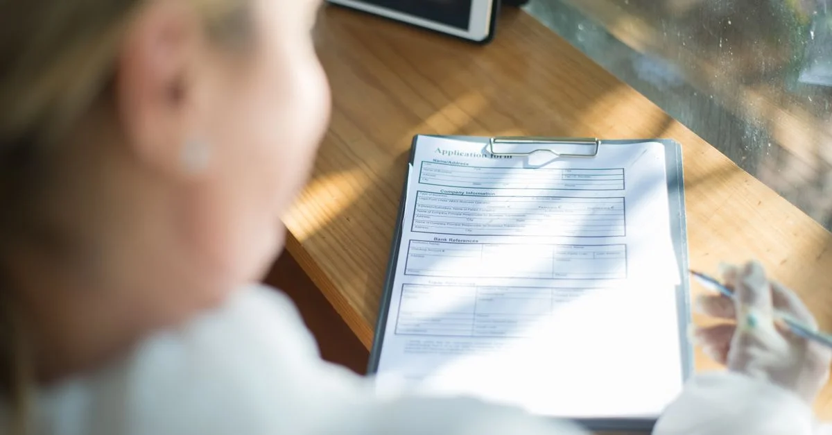 Une femme remplit un formulaire de candidature avec un stylo sur un bureau en bois baigné de lumière.