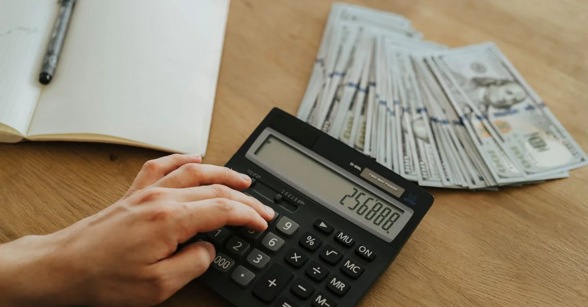 Une main calcule des chiffres financiers avec une calculatrice, entourée de piles d'argent sur une table en bois.