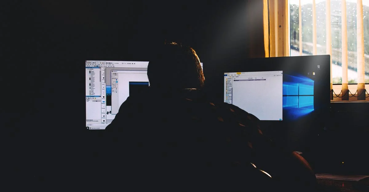 Silhouette d'une personne travaillant sur des ordinateurs dans un environnement de bureau faiblement éclairé.