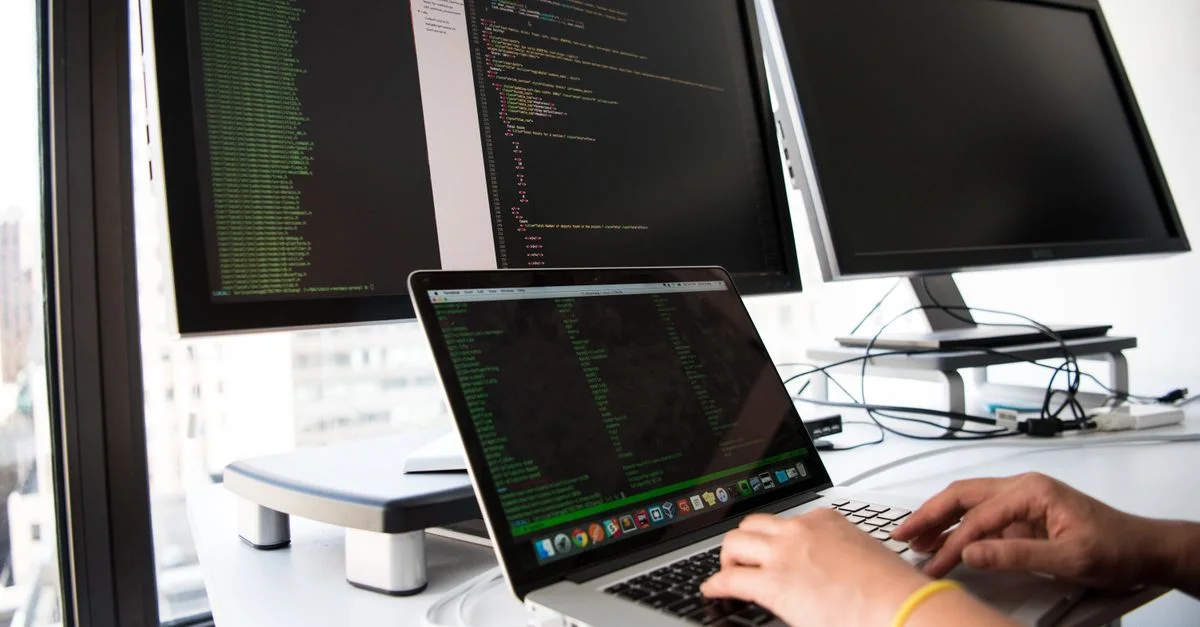 Un développeur écrit du code sur un ordinateur portable devant plusieurs moniteurs dans un bureau.