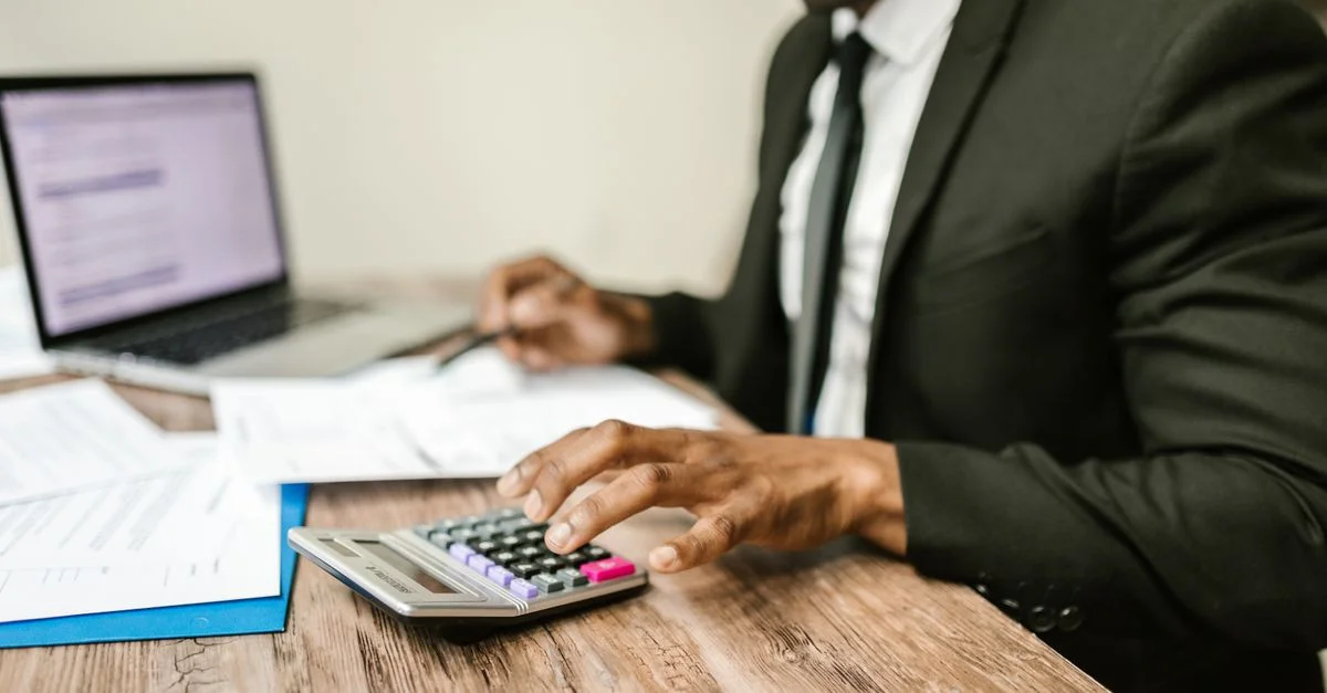 Un homme d'affaires en costume noir utilisant une calculatrice à un bureau avec des documents financiers et un ordinateur portable.