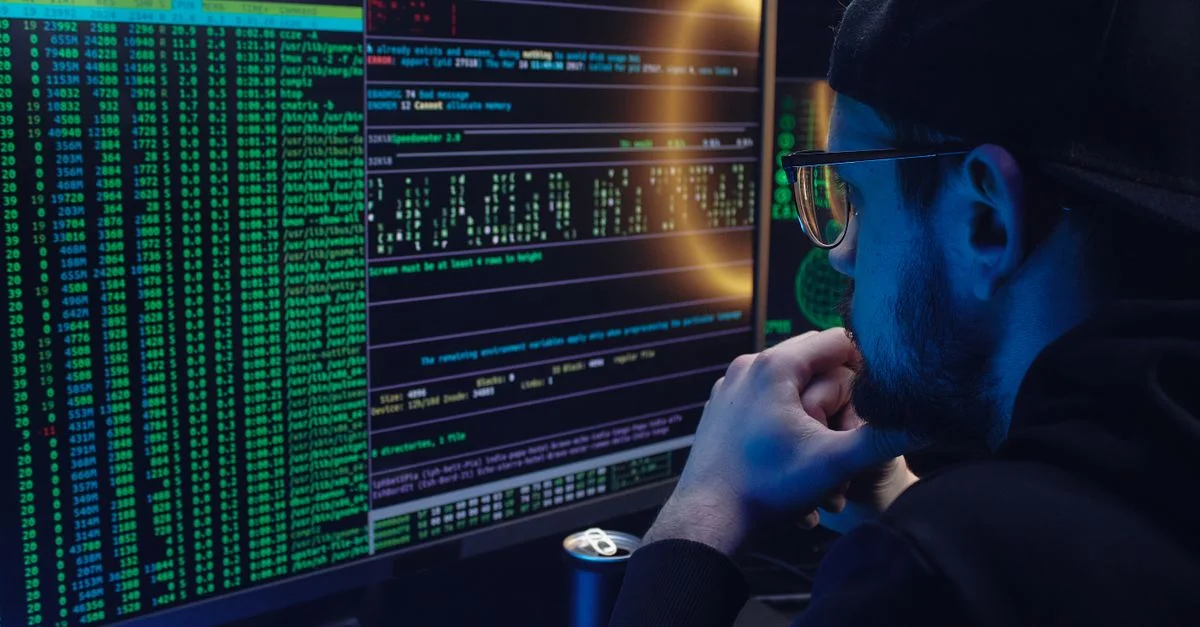Un homme travaillant intensément sur la programmation informatique avec du code affiché sur des moniteurs doubles dans une pièce faiblement éclairée.