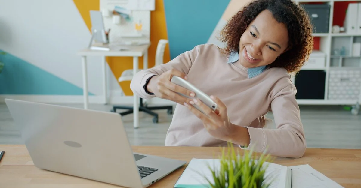 Femme souriante utilisant son smartphone et son ordinateur portable dans un espace de bureau coloré.